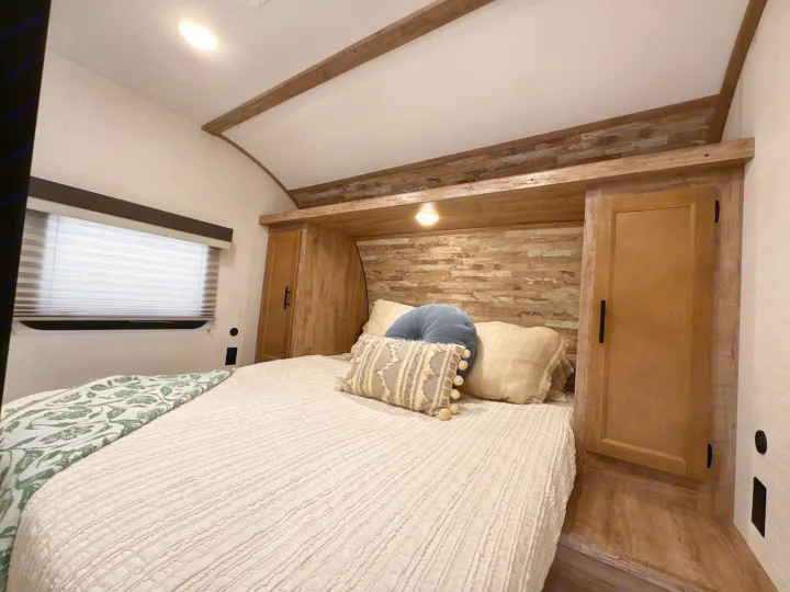 Inviting RV rental bedroom: bed with textured bedding, decorative pillows, window, and warm wood cabinetry.. Gulf Stream Enlighten 2025