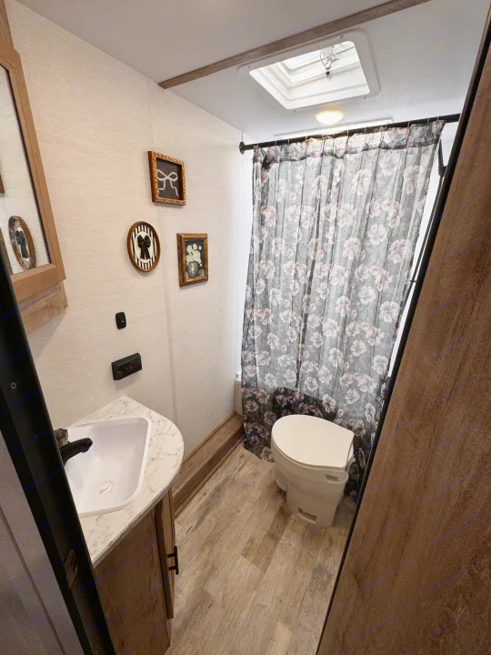 RV rental bathroom features a sink, toilet, gray floral shower curtain, and decorative wall art on light walls.. Gulf Stream Enlighten 2025