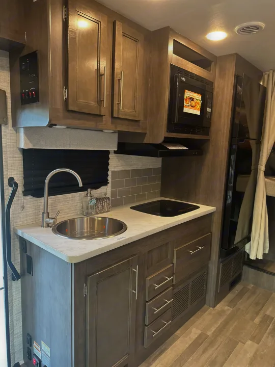 Modern RV rental kitchen with wood-grain cabinets, stainless steel sink, microwave, stove, and refrigerator. Functional and compact.. JAYCO REDHAWK SE 22TF 2025