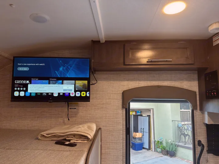 Inside an RV rental, a Connex TV displays webOS and an update prompt above a bed. A doorway leads to an outdoor patio.. JAYCO REDHAWK SE 22TF 2025