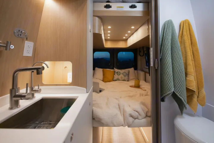 This rv rental features a compact kitchen with sink, a cozy bedroom looking out to mountains, and towels by a toilet.. Noovo Plus 2025