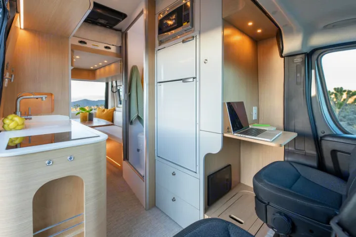 Modern RV rental interior shows kitchen, laptop workspace, and dining area with scenic outdoor views.. Noovo Plus 2025
