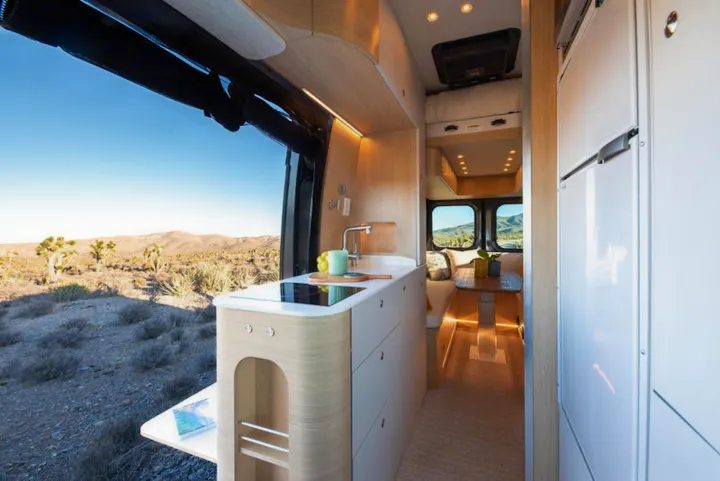 Modern RV rental interior with kitchen, dining area, and open view to a desert landscape featuring Joshua trees.. Noovo Plus 2025