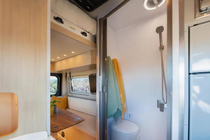 Inside an rv rental: a compact bathroom with shower & toilet, seen next to a dining area with a wooden table & overhead storage.. Noovo Plus 2025