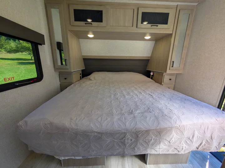 This RV rental bedroom offers a comfortable bed with a quilted cover, overhead cabinets, and a window with a green outdoor view.. Forest River Alta 2850KRL 2025