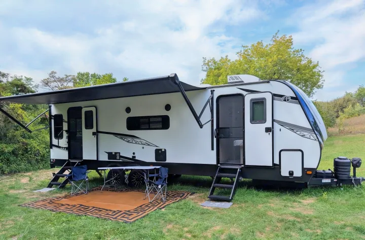 This RV rental features a white travel trailer with an extended awning, outdoor seating, and steps, parked on grass.. Forest River Alta 2850KRL 2025