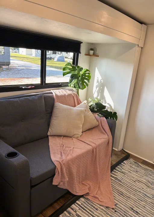 Cozy RV rental interior: A gray couch with pink blanket, Monstera plant, and window view of an RV park.. Highland Ridge RV Roamer 2017