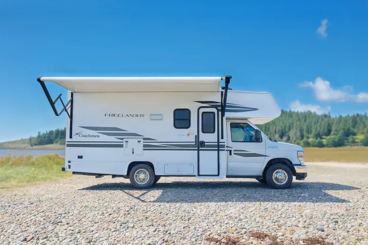 Explore scenic views! This white Coachmen Freelander rv rental, awning deployed, is parked on a gravel shoreline under a clear blue sky.. Coachman Freelander 22 XG 2023