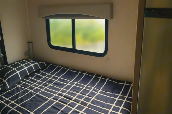 This RV rental's cozy bedroom features a bed with dark blue grid bedding under a window showing a blurry green view.. Coachman Freelander 22 XG 2023