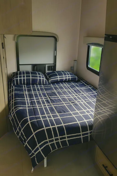 Inside an RV rental: a compact bedroom with a bed featuring dark blue & white grid bedding and two pillows. Windows let in light.. Coachman Freelander 22 XG 2023