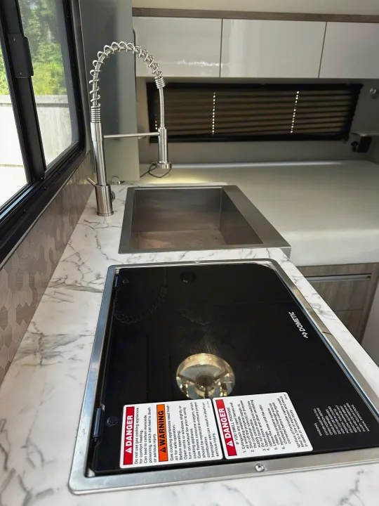 Inside a modern RV rental kitchen: a Dometic stovetop with safety warnings, a sink, and marble-look counters.. InTech RV Sol 2023