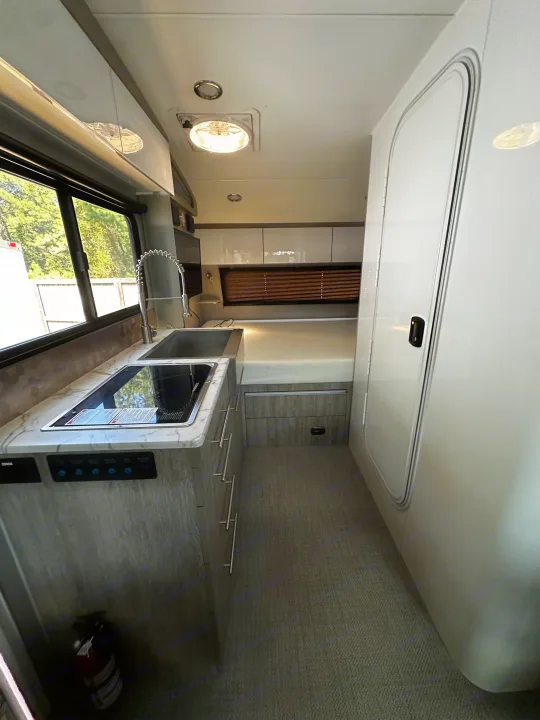 View inside an RV rental, showcasing a compact kitchenette with sink & cooktop, a bed, overhead cabinets, and floor.. InTech RV Sol 2023