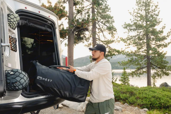 Man loads large HEST gear bag into his RV rental at a lakeside campsite, ready for van life outdoor adventure.. Airstream Basecamp 2022