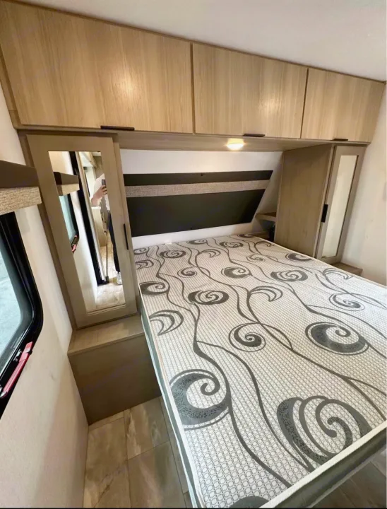 This RV rental bedroom offers a comfortable bed, ample overhead storage, and a handy mirror.. Dutchmen Kodiak 2025