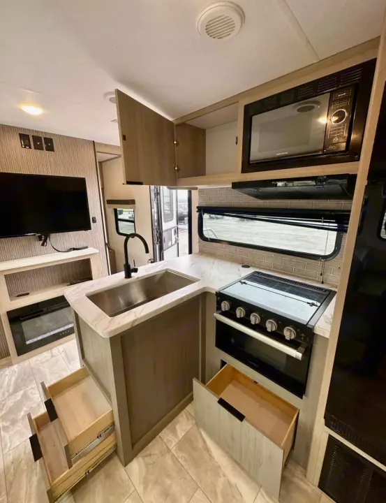 Inside this RV rental: a modern kitchen with sink, stove, microwave, open drawers, TV, and fireplace.. Dutchmen Kodiak 2025