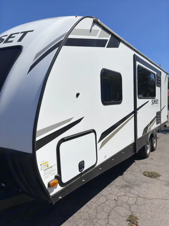 Side view of a white Sunset RV rental with black and gray graphics, parked on pavement, clear blue sky overhead.. Crossroads RV Sunset Trail Super Lite 2022