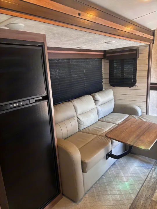 RV rental interior: Beige reclining sofa, built-in table, and refrigerator, set against modern wood trim and blinds.. Crossroads RV Sunset Trail Super Lite 2022