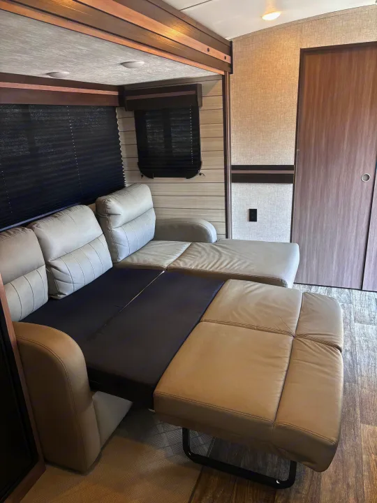 An rv rental's interior with a beige convertible sofa, partially extended into a bed, alongside windows with blinds and a door.. Crossroads RV Sunset Trail Super Lite 2022