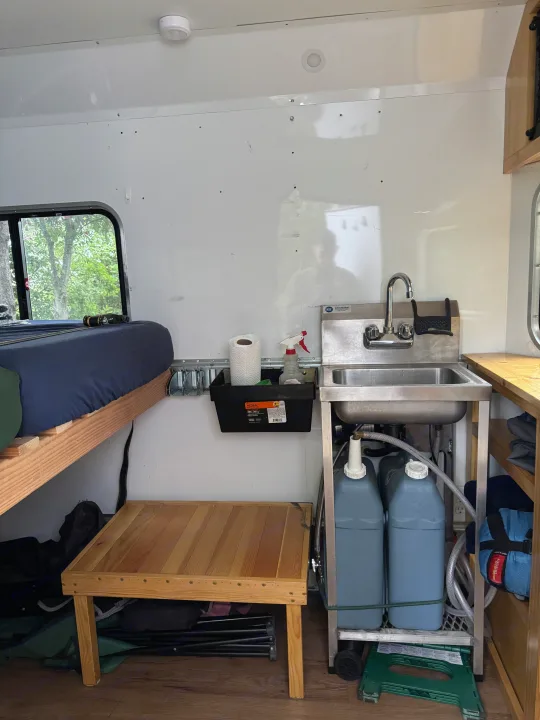 Cozy RV rental interior with a bed, wooden table, and compact kitchenette featuring a sink and water storage.. Weeroll Silver cloud 2020