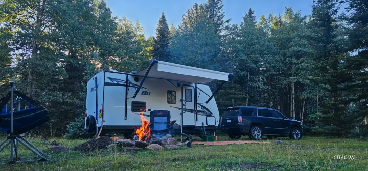 Peaceful RV rental campsite: white travel trailer, glowing campfire, and SUV nestled among tall forest trees.. Keystone RV Bullet Crossfire 2021
