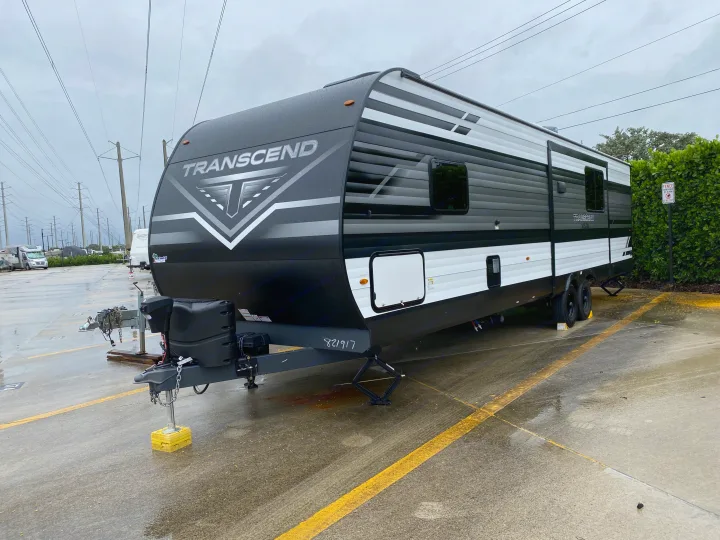 A modern black and white Transcend travel trailer, ready for RV rental, parked on a wet lot under an overcast sky.. Grand Design Transcend Xplor 2022