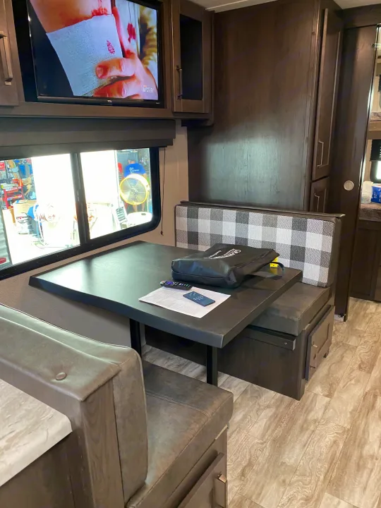 Interior of an RV rental: a modern dinette with plaid cushions, table, TV, and dark cabinetry, ready for comfortable travel.. Grand Design Transcend Xplor 2022