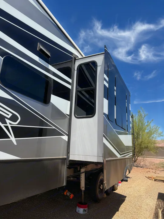 A modern RV rental with an extended slide-out is parked on gravel under a blue sky, stabilized by a jack block for travel.. Keystone RV Raptor Toy Hauler 2021