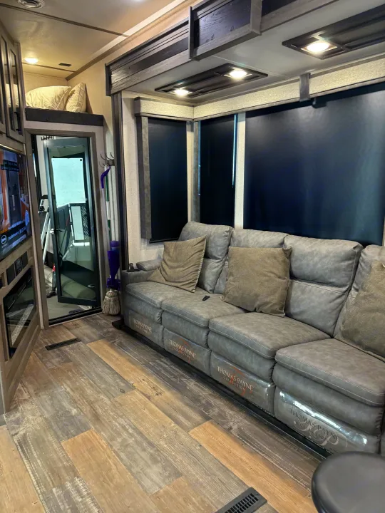 Modern RV rental living space featuring a gray "Thomas Payne Collection" sofa, wood-look floor, and dark blinds.. Keystone RV Raptor Toy Hauler 2021