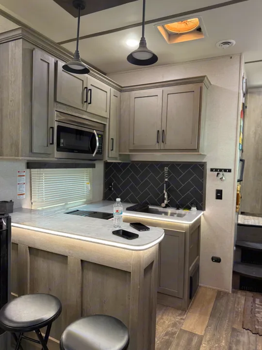Stylish RV rental kitchen: gray cabinets, black herringbone tile, island with LED lighting, microwave, sink & bar stools.. Keystone RV Raptor Toy Hauler 2021