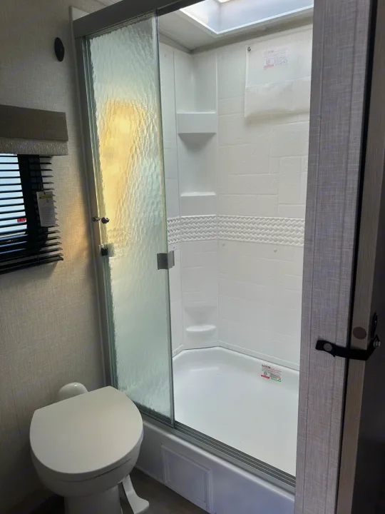 Compact RV rental bathroom with a white toilet on the left and a glass-door shower stall on the right, under a skylight.. Forest River Flagstaff Micro Lite 2023