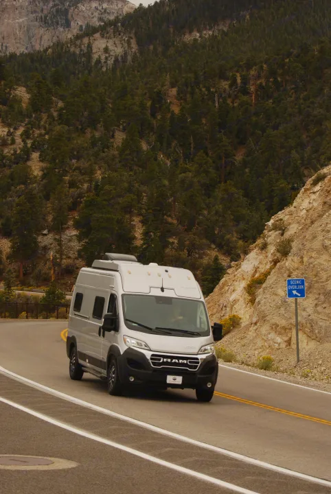 Grey RV rental driving on a scenic mountain road, lined with trees and a rocky slope, near a "Scenic Overlook" sign.. Winnebago Soilis 2025