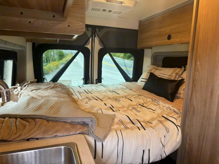 Inside a compact rv rental: cozy bed with graphic bedding, small sink, overhead cabinets, and a scenic view through rear windows.. Winnebago Soilis 2025