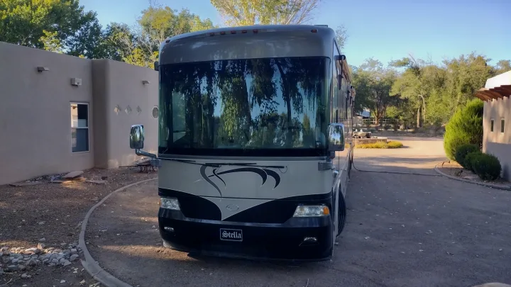 Front view of a grey and black RV rental, 'Stella,' parked on a sunny day beside a building with trees reflecting in its windshield.. Country Coach Allure 470 2005