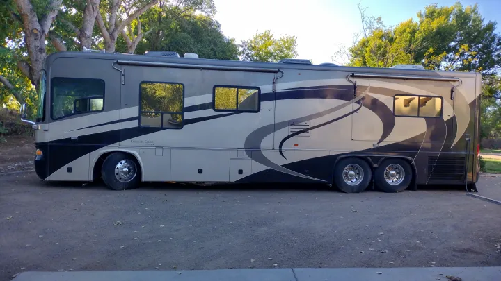 View of a large, well-maintained luxury rv rental, grey and black with stylish graphics, parked outdoors amongst trees.. Country Coach Allure 470 2005