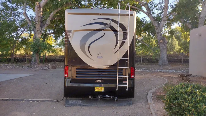 Rear view of an RV rental with a ladder, parked in a New Mexico campsite among large trees. Includes tow hitch.. Country Coach Allure 470 2005
