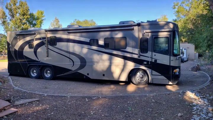 A luxurious Country Coach rv rental with a black and tan design, multiple awnings, parked amidst trees on a gravel pad.. Country Coach Allure 470 2005