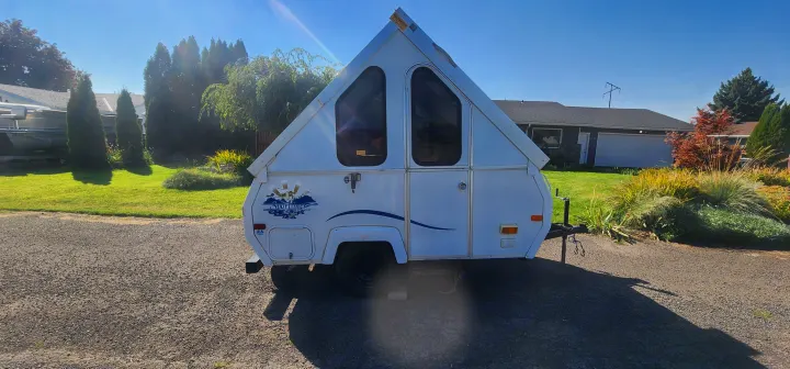White A-frame rv rental camper with triangular windows, parked on gravel in a sunny residential setting.. A Liner A Liner Popup Trailer 2006