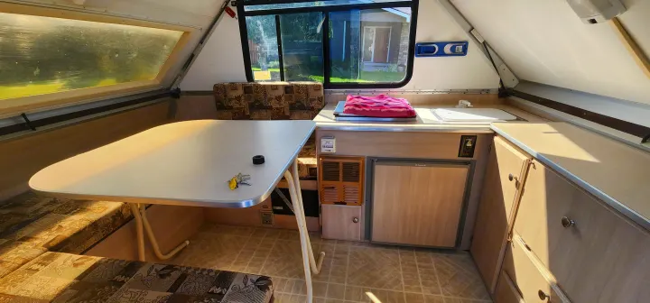 Cozy A-frame RV rental interior, featuring a dining area with table and benches, and a compact kitchenette with sink, stove, and fridge.. A Liner A Liner Popup Trailer 2006