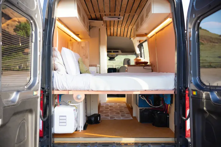 Rear doors open to a custom rv rental camper's cozy interior with a bed, wood ceiling, and off-grid utilities.. Ford Transit 2023