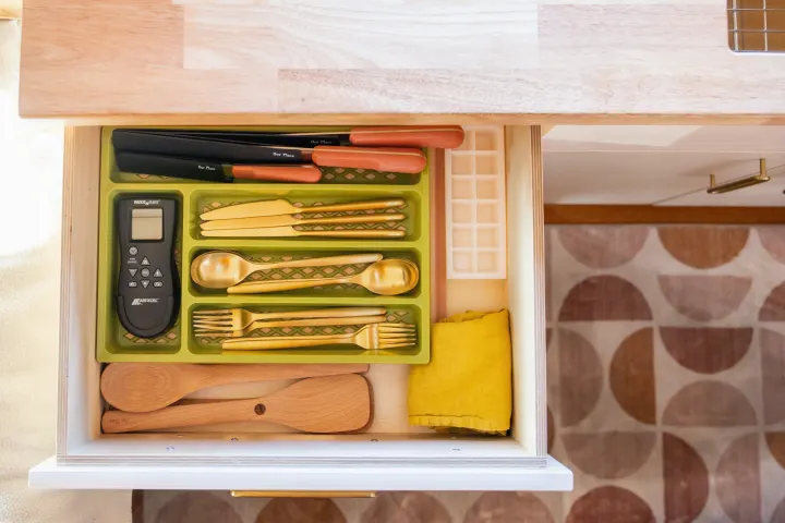 Organized RV rental kitchen drawer with golden cutlery, MaxxAir remote, Our Place knives, and wooden utensils.. Ford Transit 2023