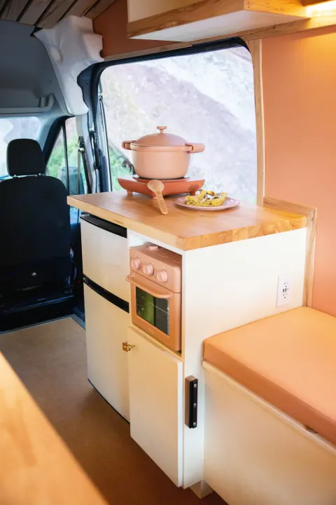 RV rental kitchen: a chic salmon-pink pot on an induction cooktop, oven, fridge, and cozy bench. Van life essentials.. Ford Transit 2023