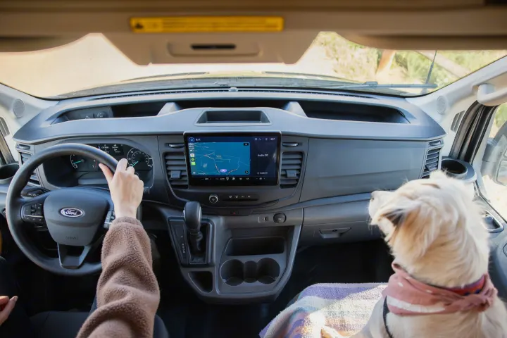RV rental interior: A person drives a Ford van with a dog co-pilot, viewing navigation on the infotainment screen.. Ford Transit 2023