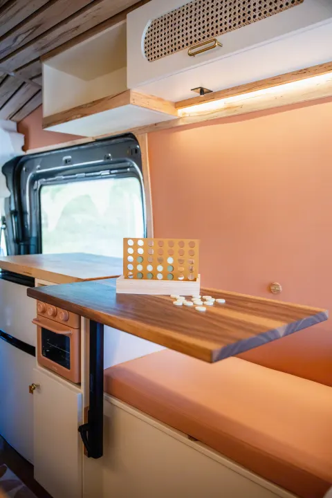 This inviting RV rental interior features a Connect Four game on a wooden table, warm peach walls, and custom wood cabinetry.. Ford Transit 2023