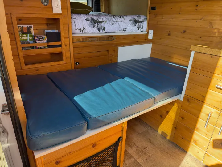 Cozy RV rental features wood walls, blue convertible seating, an overhead bunk bed, and storage for van life adventures.. RAM Promaster 2500 2020