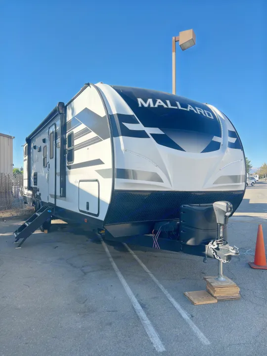 Mallard travel trailer RV rental parked on asphalt, front hitch on wood blocks, with steps extended under a clear blue sky.. Heartland RVs Mallard 2022