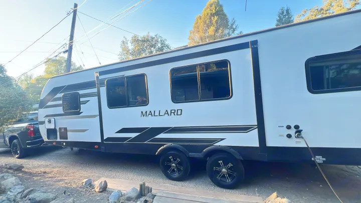 This white Mallard travel trailer rv rental is parked outdoors, hitched to a truck. Power is connected; a person waves from inside.. Heartland RVs Mallard 2022