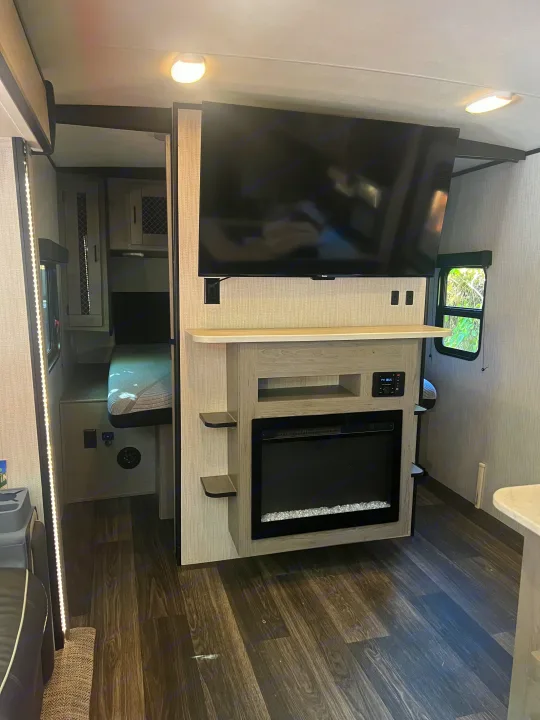 Modern **rv rental** living area with a large TV, electric fireplace, and cozy bunk beds. Dark wood floors and bright lighting enhance comfort.. Heartland RVs Mallard 2022