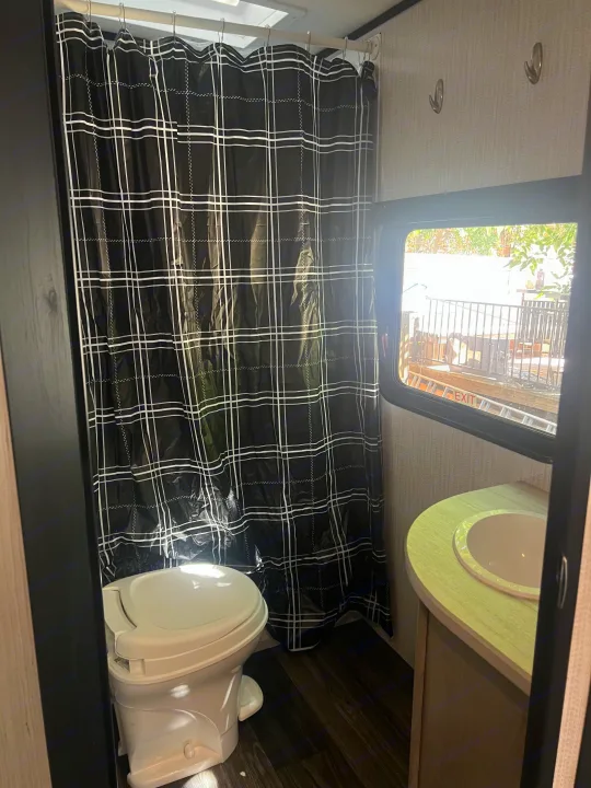 Enjoy this compact RV rental bathroom with a black plaid shower curtain, toilet, and sink, offering natural light and an outdoor view.. Heartland RVs Mallard 2022
