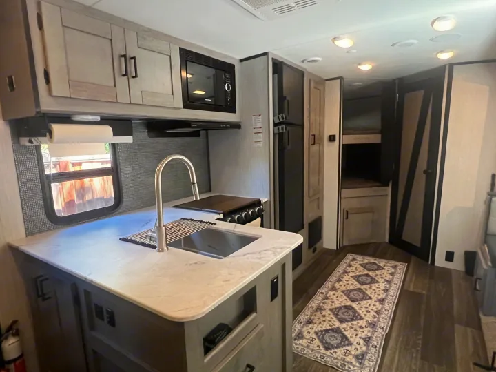 Modern RV rental kitchen: sink, counter, microwave, fridge, bunk beds & floor rug, lit by overhead lights.. Heartland RVs Mallard 2022