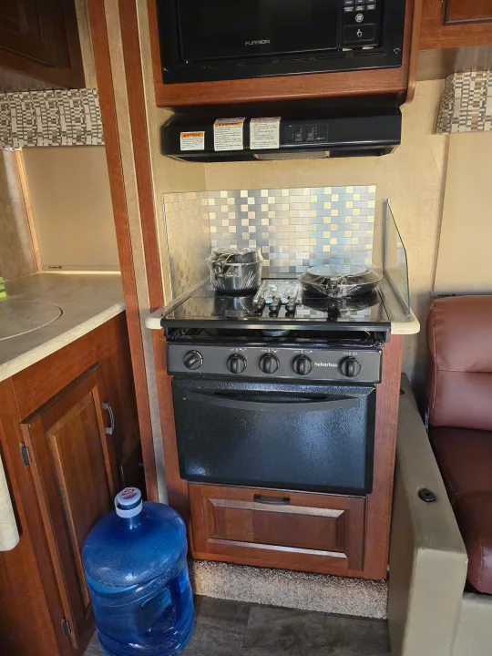 This rv rental kitchen features a microwave, Suburban stove/oven, metallic backsplash, and a large water bottle on the floor.. Forest River Sunseeker 2016